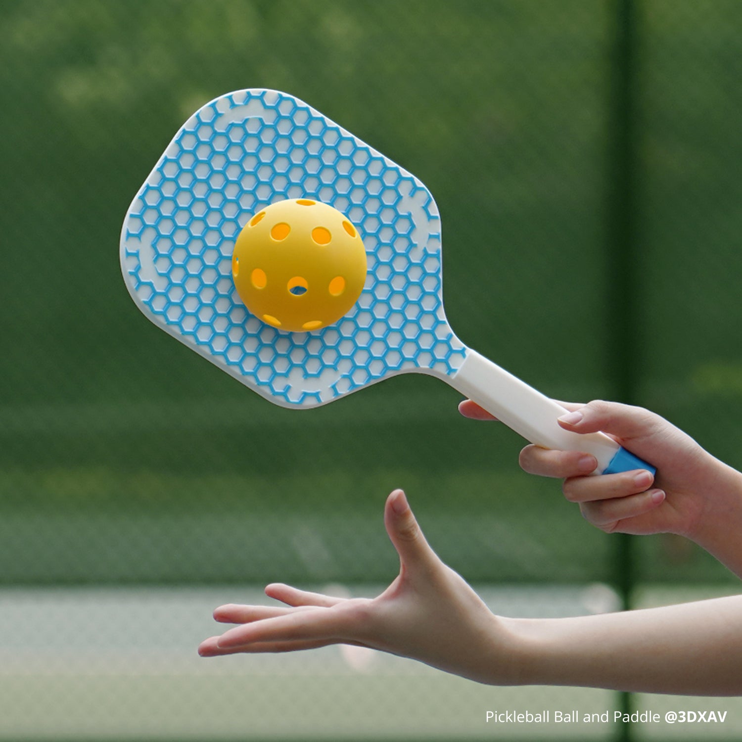 PLA TOUGH+ FIlament Pickleball paddle with a yellow ball held by a hand against a green outdoor background